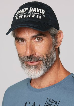 Navy baseball cap with white embroidered text "CAMP DAVID" and "BLUE CREW 63." Light blue crew neck t-shirt with black graphic print.