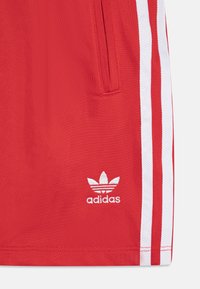 Červené športové šortky s bielymi bočnými pruhmi a vyšívaným logom Adidas v dolnom ľavom rohu. Vyrobené z ľahkej, štruktúrovanej tkaniny.