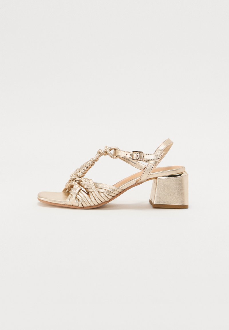 Gold heeled sandal featuring crisscrossed straps, a braided detail, and a square block heel. Smooth texture with metallic finish.