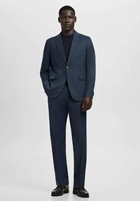 Mężczyzna stojący z rękami w kieszeniach, ubrany w ciemnoniebieski garnitur na miarę, czarną koszulę i czarne eleganckie buty na jednolitym szarym tle.
