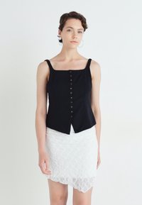 Svart avkortet tanktop med firkantet halslinje, med frontknapper og en utvidet kant. Kombinert med en hvit blondet omslagsskjørt.