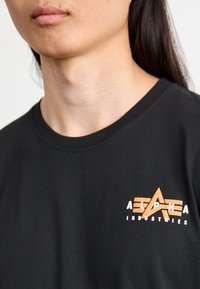 Crna pamučna majica s logotipom na kojem je narandžasti i bijeli tekst "ALPHA INDUSTRIES" uz grafiku zvijezde. Kratki rukavi.