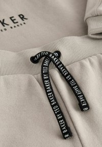 Pantalons jogger beige clair avec une taille côtelée et un cordon noir avec une marque répétée. Texture douce et design décontracté.