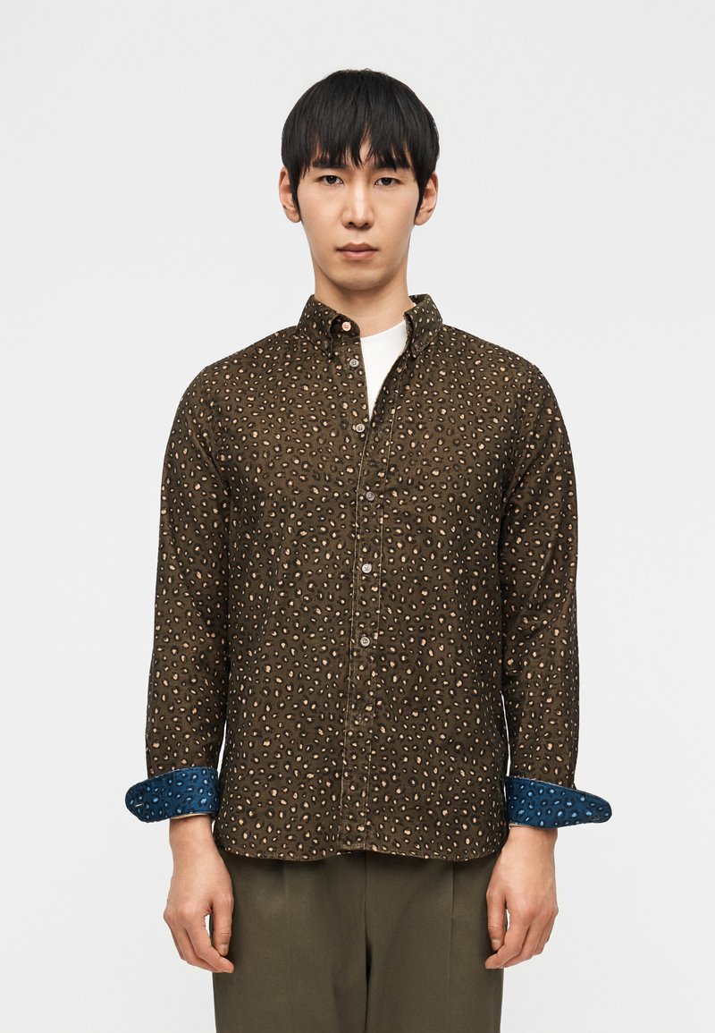 Άνδρας που φοράει καφέ πουκάμισο με κουμπιά, με ανοιχτόχρωμες κηλίδες και μανσέτες με μπλε μοτίβα, στέκεται μπροστά σε απλό λευκό φόντο.