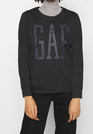 Personne portant un sweat-shirt noir avec le logo "GAP" sur un col roulé rayé noir et blanc et un pantalon noir.