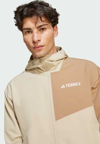 Beige capuchonjack met een contrasterend bruin paneel en een wit logo. Gladde textuur, volledige ritssluiting en lichtgewicht ontwerp.