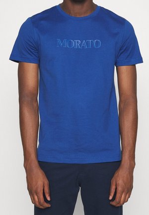 Homme portant un T-shirt ajusté bleu royal avec le texte « MORATO » en bleu clair sur la poitrine, associé à un pantalon foncé.