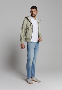 Lichtgroene hooded jas met ritssluitingen, witte polo, blauwe jeans en grijze sneakers met witte strepen. Soepel, casual ontwerp.