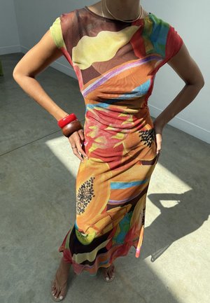 Persona che indossa un vestito maxi colorato e aderente con motivi astratti e frutta, in piedi con le mani sui fianchi, con braccialetti rossi e marroni.