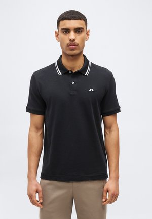 Férfi fekete galléros pólóban, fehér szegéllyel a galléron és bézs nadrágban, világos, egyszínű háttér előtt állva.