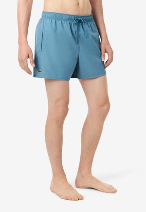 Homme portant un short de bain bleu avec taille élastique et cordon, debout pieds nus sur fond blanc.