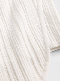 Texturovaná bílá látka s vlnitým vzorem, která má jemné rýhy a hladké okraje. Materiál vypadá hebce a lákavě.