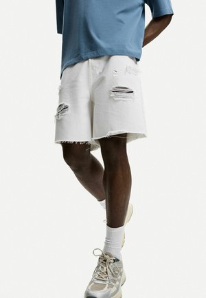White denim shorts with frayed hem and multiple rips. Worn with a blue T-shirt and beige sneakers featuring mesh and rubber accents.