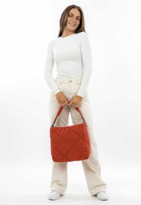 Quilted red handbag with a soft texture, rectangular shape, and short strap, featured in a minimalistic setting. White top and light pants worn.