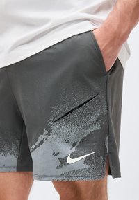 Άτομο που φορά σκούρα γκρι σορτς Nike με λευκό λογότυπο Swoosh και σχέδιο πιτσιλιών χρώματος, το χέρι τοποθετημένο στην δεξιά τσέπη.