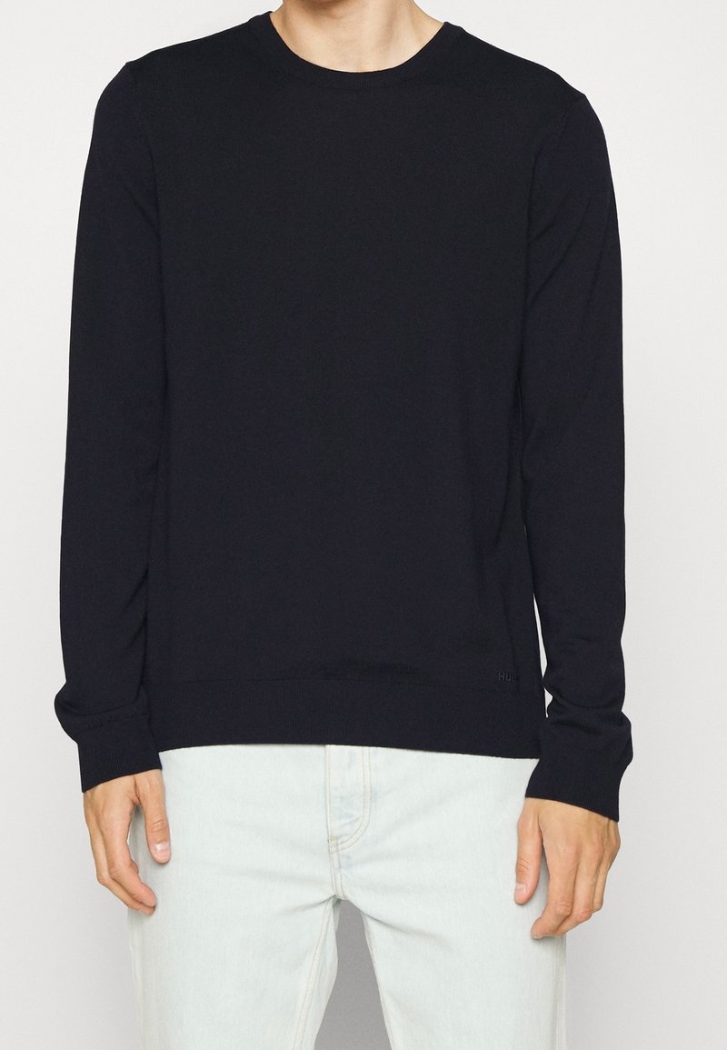 Personne portant un pull noir uni à manches longues et un pantalon clair, debout devant un fond uni.