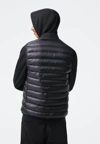 Svart puffervest med horisontal quilt, skinnende tekstur og høy krage, båret over en svart hettegenser, sett fra baksiden.