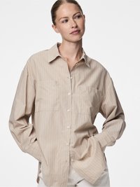 Lengva smėlio spalvos marškiniai su baltomis vertikaliomis juostelėmis, dviem priekinėmis kišenėmis ir laisvu siluetu. Pagaminti iš minkštos, tekstūruotos medžiagos.