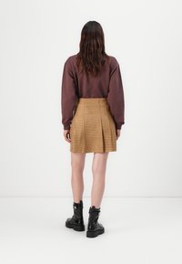 Hnědá oversized mikina s dlouhými rukávy, spárovaná se žlutou kostkovanou plisovanou sukní a černými kotníkovými botami. Bílý pozadí.
