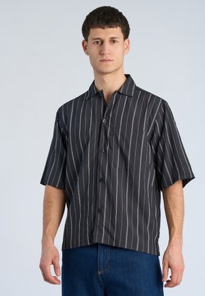 Uomo che indossa una camicia nera a maniche corte con righe verticali bianche e jeans blu, in piedi davanti a uno sfondo chiaro e uniforme.
