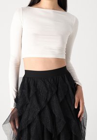 Femme portant un crop top blanc à manches longues et une jupe en tulle noire superposée avec des détails scintillants, les mains touchant délicatement la jupe.