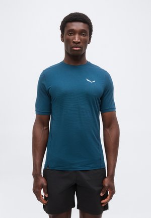 Man die recht vooruit kijkt, draagt een teal kleurig sportshirt met korte mouwen en zwarte korte broek tegen een effen witte achtergrond.