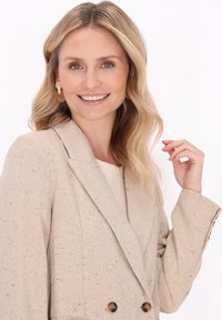 Blazer doppiopetto beige con texture maculata in vari colori e bottoni marroni, indossato sopra un top chiaro.