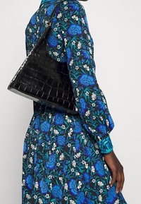 Borsa triangolare nera in pelle effetto coccodrillo con una sola tracolla, indossata su un vestito a stampa floreale con colori blu e bianchi su uno sfondo nero.