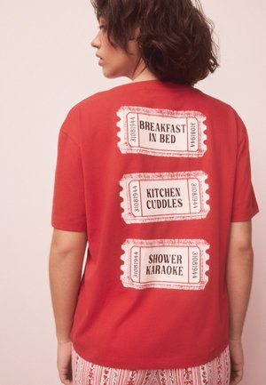 Henkilö pukeutuneena punaiseen t-paitaan, jonka takana on valkoisia lippusuunnitteluita, joissa lukee "Aamiainen sängyssä", "Keittiöhalaus" ja "Suihku karaokea".