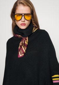 Μαύρο πλεχτό πουλόβερ με μανίκια τσέρι και με κασκόλ από μετάξι σε χρώμα μπορντό με σχέδια. Οι υπερμεγέθεις μαύρες γυαλιά με κίτρινους φακούς ολοκληρώνουν το σύνολο.