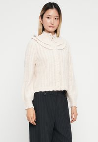 Lys rosa strikket cardigan med volangkrage, knappelukking og ribbestrikket tekstur, kombinert med mørke stripete bukser.
