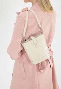Lys beige spandbags fremstillet af tekstureret læder, med en aftagelig rem og et håndtag i toppen, hængende på en skulder.