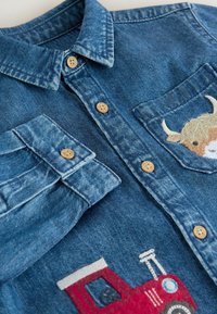 Camicia di denim con colletto classico, bottoni in legno e dettagli ricamati di un trattore e di una mucca sulle tasche frontali.