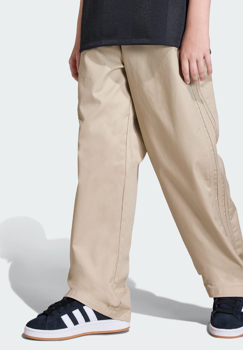 Beżowe, luźne spodnie wykonane z lekkiego materiału, z pionowymi szwami i szerokimi nogawkami. Połączone z granatowymi sneakersami z białymi paskami.