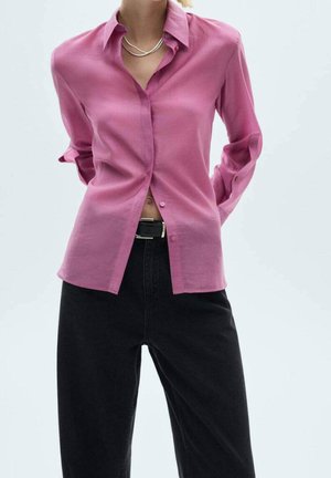 Kvinde iført en pink skjorte med knapper over sorte bukser med høj talje og sort bælte, stående med hænderne bag ryggen.