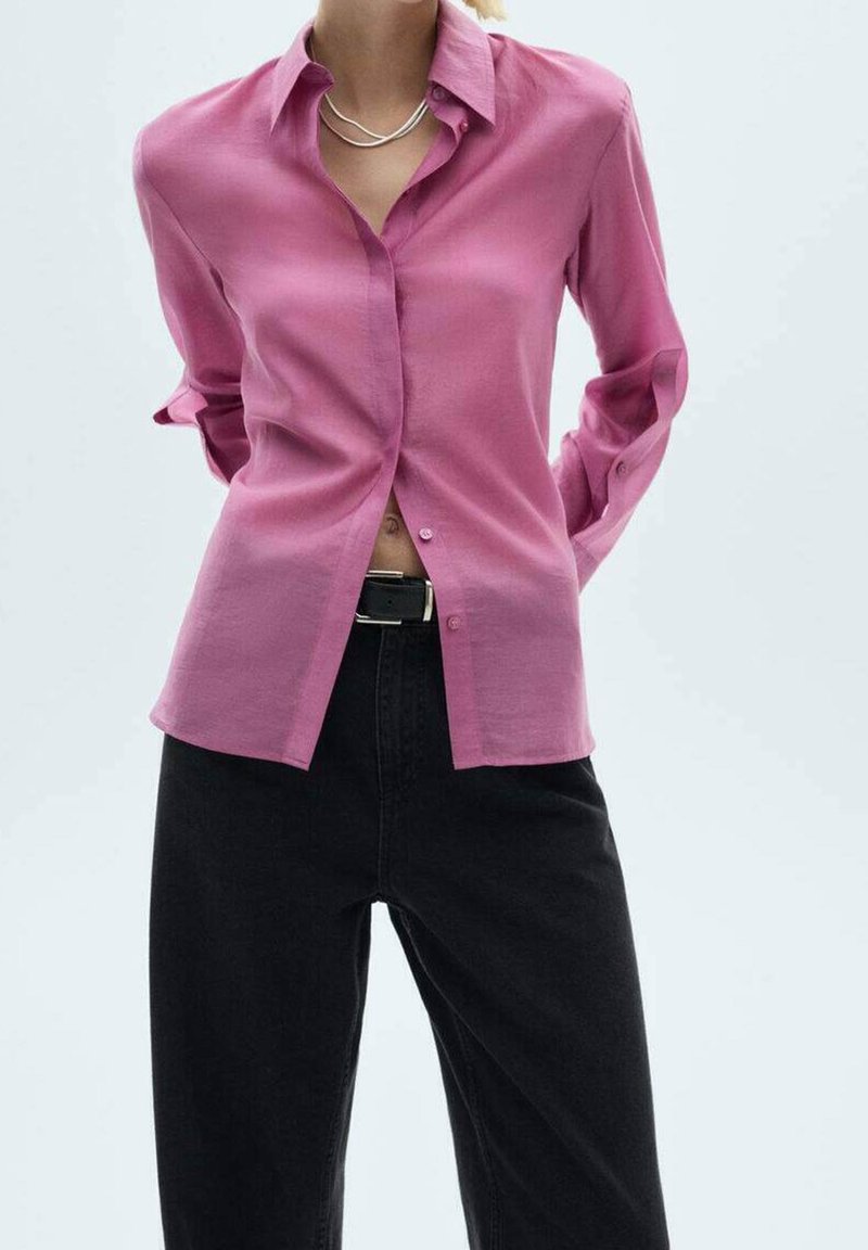 Femme portant une chemise rose boutonnée sur un pantalon noir taille haute avec une ceinture noire, debout les mains derrière le dos.