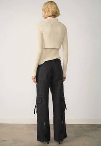 Rebrasti kremasti pulover s turtleneckom i skraćenim leđima, u kombinaciji s crnim širokim cargo hlačama koje imaju zatvarače na rubovima i bočne džepove.