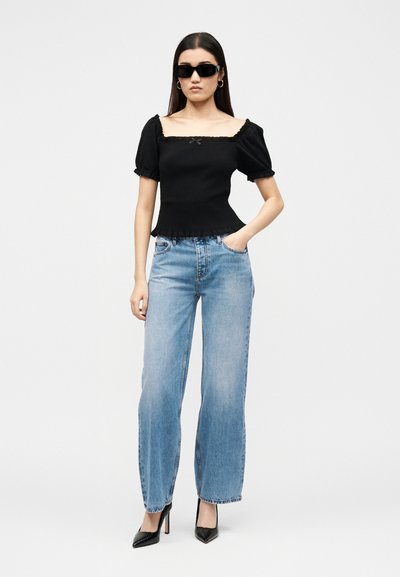 Kvinne iført svart topp med pufferm, lyseblå vide jeans, svarte spisse høye hæler og svarte solbriller, stående med den ene hånden i lommen.