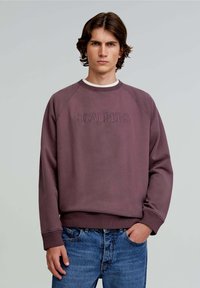 Felpa bordeaux con polsini e orlo a coste, con logo "SCALPERS" in rilievo sul davanti. Abbinata a jeans in denim blu.
