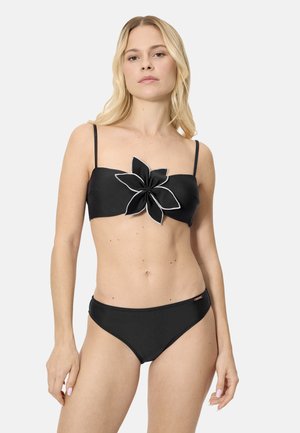 Mujer rubia con un bikini negro de tirantes finos y un gran detalle de flor en la parte superior tipo bandeau, posando frente a un fondo liso.
