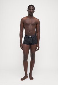 Fekete boxer alsónadrág rugalmas anyagból, geometrikus mintával és derékpánttal. A modell áll, mezítláb, egy letisztult háttér előtt.