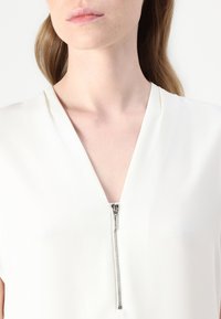 Blusa blanca con escote en V y mangas cortas, que presenta una cremallera plateada en el centro delantero, hecha de una tela suave y ligera.