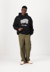Hoodie preto com impressão gráfica, calças cargo verde-oliva e sapatilhas claras com riscas vermelhas. Modelo está de pé diante de um fundo branco simples.