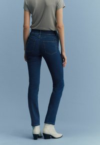 Donkerblauwe denim jeans met een slim fit, voorzien van twee achterzakken en een medium taille. Het model draagt ze met crèmekleurige enkellaarsjes.