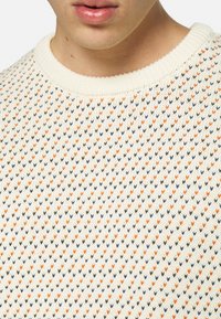 Krämfärgad stickad tröja med ett strukturerat mönster av små hjärtformer i orange, blått, grönt och svart. Rund halsringning.