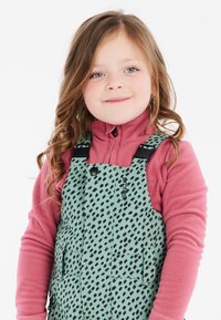 Bambino che indossa una salopette verde menta con un motivo a diamond nero sopra una maglietta di pile rosa. La salopette ha tasche funzionali e bretelle.