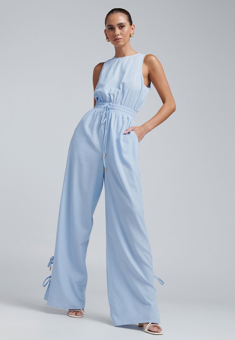 Gaiši zils kombinezons ar pieguļošu vidukli, platām kāju daļām un regulējamiem auklu elementiem pie potītēm. Gluda auduma ar tīru kakla izgriezumu.