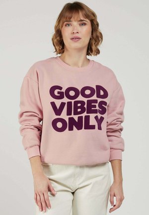 Femme aux cheveux courts et ondulés portant un sweat-shirt rose avec l'inscription "GOOD VIBES ONLY" en lettres majuscules violettes, associé à un pantalon de couleur claire.