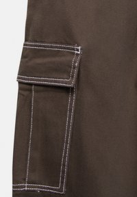 Brun tygficka med kontrasterande vit sömnad, med en klaffstängning och en strukturerad yta, som är en del av ett större plagg.