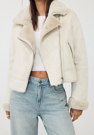 Femme portant une veste en peau lainée couleur crème avec des poignets repliés, un crop top blanc et un jean taille haute bleu clair.